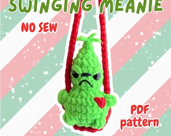Op de afbeelding: Een groene gehaakte Grinch-achtige karakter met een rood hart, hangend aan rode koorden. De tekst "SWINGING MEANIE" en "NO SEW" staan boven het karakter. De tekst "PDF pattern" en "@CRAFTYBAKERDESIGNS" zijn ook zichtbaar.