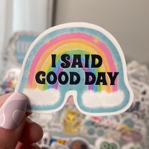 Op de afbeelding: Een sticker met een regenboog en wolken, met de tekst "I SAID GOOD DAY" in zwart.