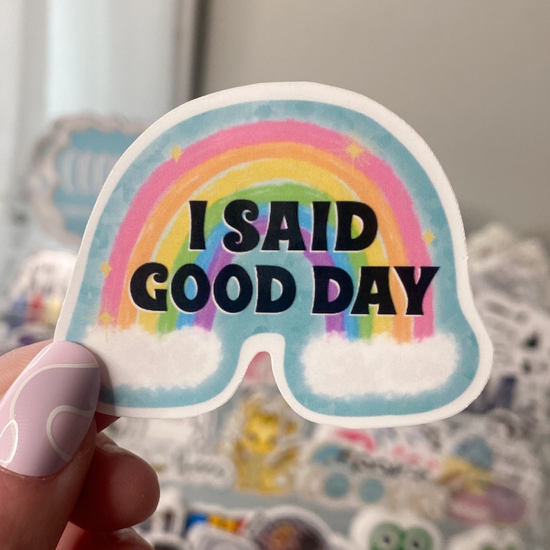 Op de afbeelding: Een sticker met een regenboog en wolken, met de tekst "I SAID GOOD DAY" in zwart.