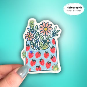 以下が含まれることがあります： 赤いイチゴ、花、緑のカエルが描かれた青いバッグのデザインのホログラフィックビニールステッカー。「Holographic Vinyl Sticker」の文字が白い楕円で囲まれています。