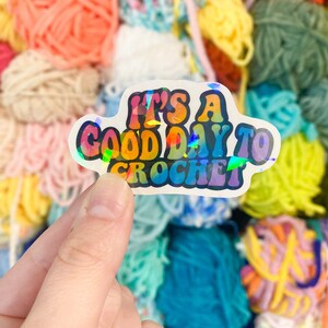 May include: A holographic sticker with the text "It's a good day to crochet" in colorful, retro-style lettering. The sticker is held in a hand against a backdrop of colorful yarn skeins in various shades of pink, blue, yellow, and green.