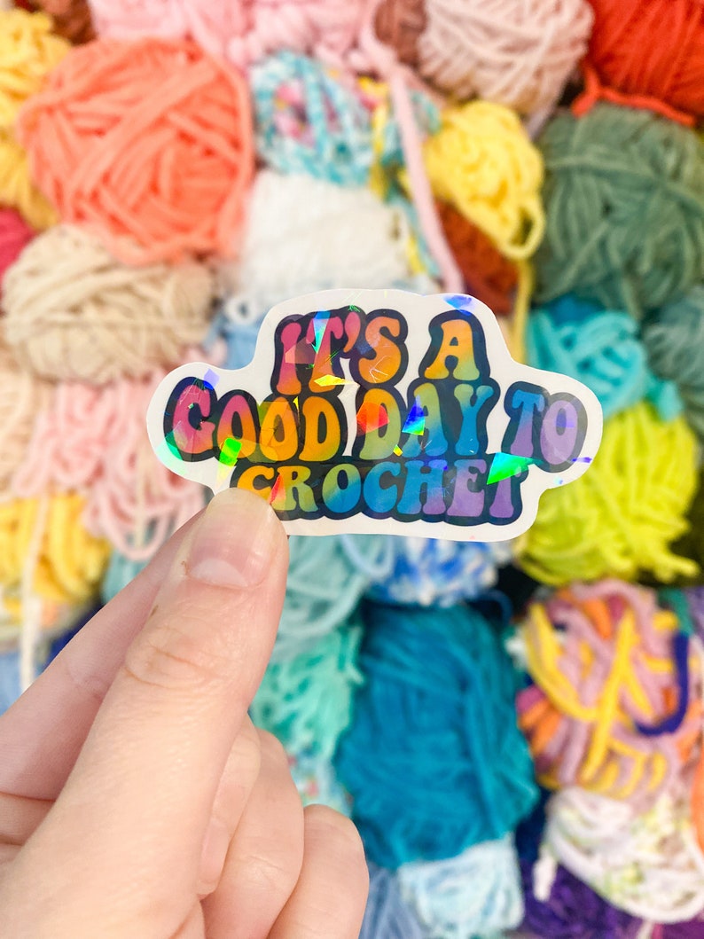 May include: A holographic sticker with the text "It's a good day to crochet" in colorful, retro-style lettering. The sticker is held in a hand against a backdrop of colorful yarn skeins in various shades of pink, blue, yellow, and green.