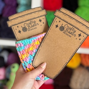 May include: A handmade, colorful crochet coffee cup sleeve held with two brown cardboard coffee cup-shaped tags. The tags have the text "Crafty Baker Designs" and a logo. The sleeve is a mix of blue, yellow, pink, and orange yarn.