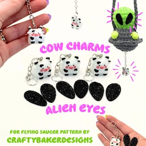 May include: Small white cow charms with black spots and pink cheeks attached to silver keychains, alongside black glittery teardrop-shaped alien eyes. The text reads COW CHARMS, ALIEN EYES, FOR FLYING SAUCER PATTERN BY CRAFTYBAKERDESIGNS.