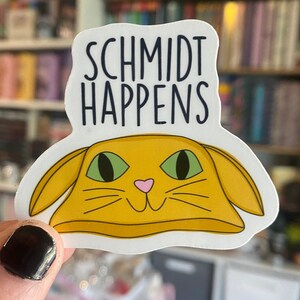 以下が含まれることがあります： 黒い文字で「Schmidt Happens」と書かれた白いステッカー。テキストの下には、緑色の目とピンク色の鼻をした黄色の猫の頭が描かれています。