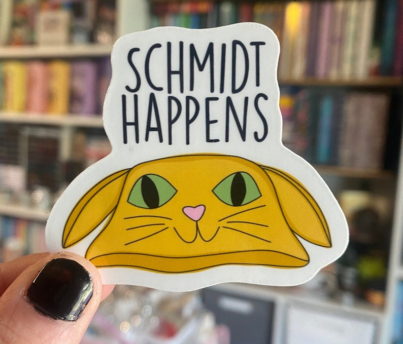 以下が含まれることがあります： 黒い文字で「Schmidt Happens」と書かれた白いステッカー。テキストの下には、緑色の目とピンク色の鼻をした黄色の猫の頭が描かれています。