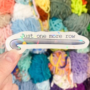 May include: A white sticker with a holographic sheen featuring a crochet hook design and the text "Just one more row" in green and black. The sticker is held in front of a colorful background of yarn balls in various colors.