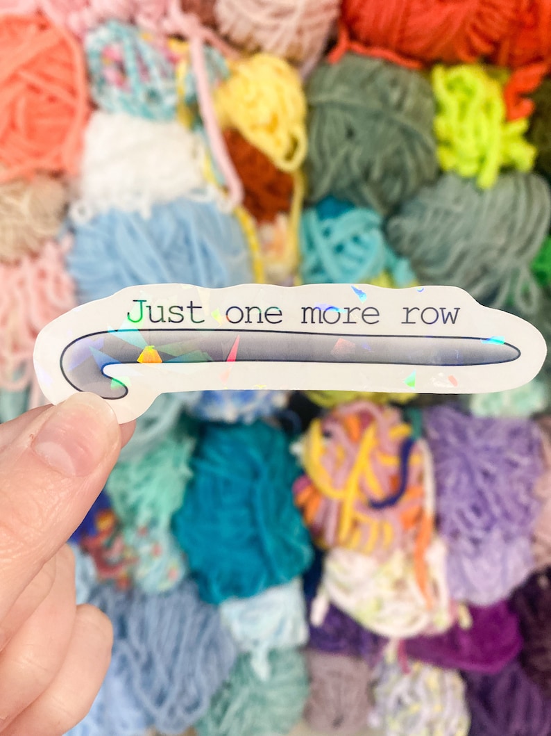 May include: A white sticker with a holographic sheen featuring a crochet hook design and the text "Just one more row" in green and black. The sticker is held in front of a colorful background of yarn balls in various colors.