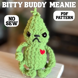 May include: A green crocheted doll with black eyes, a black frown, and a red heart. The doll is labeled "Bitty Buddy Meanie" and "PDF Pattern". The words "No Sew" are also visible.