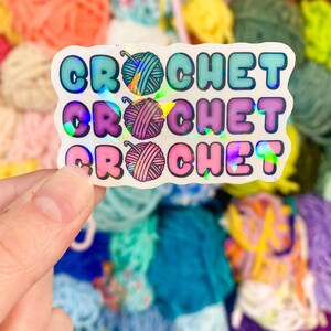 May include: A holographic sticker with the word "CROCHET" repeated three times in different colors. Each word has a small illustration of a yarn ball. The background is a colorful assortment of yarn skeins.