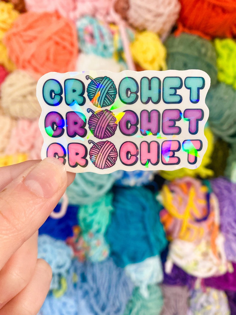 May include: A holographic sticker with the word "CROCHET" repeated three times in different colors. Each word has a small illustration of a yarn ball. The background is a colorful assortment of yarn skeins.