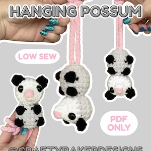 Op de afbeelding: Gehaakte hangende opossum-speeltjes in wit, zwart en roze. De speeltjes hebben zwarte oren en vlekken, roze neuzen en worden opgehangen aan roze koorden. De afbeelding bevat de tekst "HANGING POSSUM", "LOW SEW", "PDF ONLY" en "@CRAFTYBAKERDESIGNS".