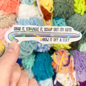 May include: A holographic sticker in the shape of a crochet hook, held in a hand, with the text "GRAB IT, STRANGLE IT, SCOOP OUT ITS GUTS, THROW IT OFF A CLIFF" in black. The background is a colorful assortment of yarn.