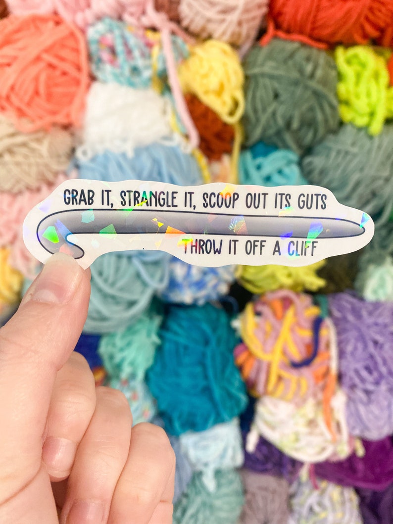 May include: A holographic sticker in the shape of a crochet hook, held in a hand, with the text "GRAB IT, STRANGLE IT, SCOOP OUT ITS GUTS, THROW IT OFF A CLIFF" in black. The background is a colorful assortment of yarn.
