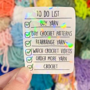 May include: A holographic sticker with a "TO DO LIST" for crochet enthusiasts. The list includes "BUY YARN", "BUY CROCHET PATTERNS", "REARRANGE YARN", "WATCH CROCHET VIDEOS", "ORDER MORE YARN", and "CROCHET". The background features colorful yarn.
