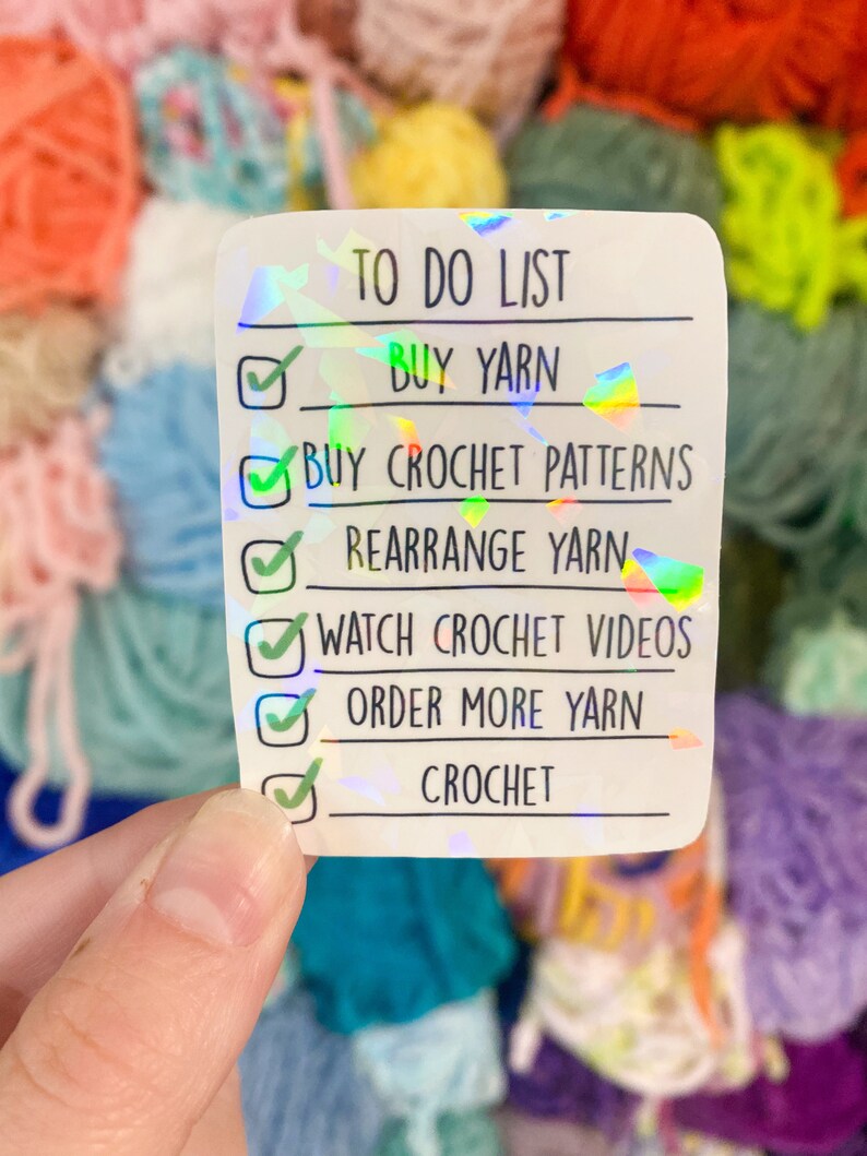 May include: A holographic sticker with a "TO DO LIST" for crochet enthusiasts. The list includes "BUY YARN", "BUY CROCHET PATTERNS", "REARRANGE YARN", "WATCH CROCHET VIDEOS", "ORDER MORE YARN", and "CROCHET". The background features colorful yarn.