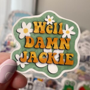 Op de afbeelding: Een groene sticker met witte madeliefjes en gele tekst die "Well Damn Jackie" luidt.