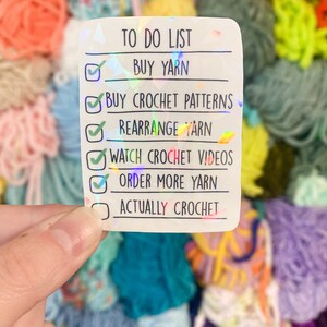 May include: A holographic sticker with a to-do list for crocheting. The list includes buying yarn, crochet patterns, rearranging yarn, watching videos, and ordering more yarn. The background features colorful yarn.
