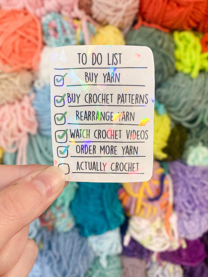 May include: A holographic sticker with a to-do list for crocheting. The list includes buying yarn, crochet patterns, rearranging yarn, watching videos, and ordering more yarn. The background features colorful yarn.