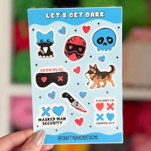 Puede incluir: Una hoja de pegatinas azul claro con varios gr&aacute;ficos de dibujos animados y texto. Los dise&ntilde;os incluyen una calavera azul, un casco rojo, un gato negro, un pastor alem&aacute;n, corazones y un cuchillo. El texto incluye "LET'S GET DARK", "LET'S PLAY", "MASKED MAN SECURITY" y "LIGHTS OUT".