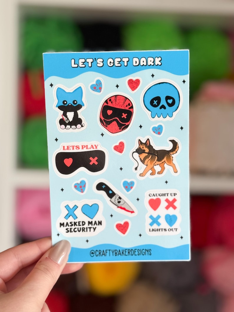 Puede incluir: Una hoja de pegatinas azul claro con varios gr&aacute;ficos de dibujos animados y texto. Los dise&ntilde;os incluyen una calavera azul, un casco rojo, un gato negro, un pastor alem&aacute;n, corazones y un cuchillo. El texto incluye "LET'S GET DARK", "LET'S PLAY", "MASKED MAN SECURITY" y "LIGHTS OUT".