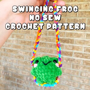 May include: A green crocheted frog with a smiling face is hanging from a colorful crocheted rope. The rope is made of multiple colors, including purple, orange, yellow, and blue. The text "Swinging Frog No Sew Crochet Pattern" is written in white letters on a black background.