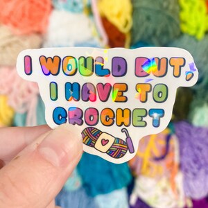 May include: A holographic sticker with the text "I WOULD BUT I HAVE TO CROCHET" in colorful bubble letters. The sticker features an illustration of yarn and a crochet hook. The background is a collection of colorful yarn balls.