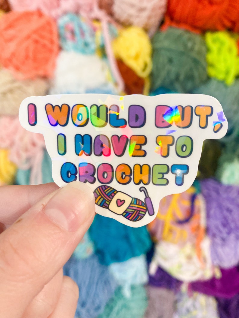 May include: A holographic sticker with the text "I WOULD BUT I HAVE TO CROCHET" in colorful bubble letters. The sticker features an illustration of yarn and a crochet hook. The background is a collection of colorful yarn balls.