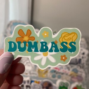 Op de afbeelding: Een groene sticker met het woord "DUMBASS" in turquoise letters met een retro lettertype. De sticker is versierd met oranje en gele bloemen.