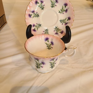 Op de afbeelding: Een vintage theekop en schotel set. De kop en schotel zijn wit met een roze rand en gouden rand. Paarse distel bloemmotieven zijn verspreid over de set. De theekop heeft een gebogen handvat.