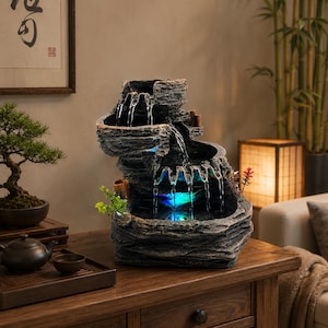 May include: A tiered tabletop water fountain with a dark, textured rock-like finish. Water cascades down three levels into a basin illuminated by a blue light. Small green artificial plants and bamboo-style accents are attached to the structure.