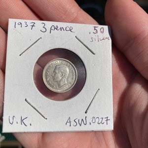 Może przedstawiać: Srebrna moneta 3-pencowa z 1937 roku z Wielkiej Brytanii. Moneta znajduje się w białym uchwycie na monety z napisem "1937 3 pence .50 silver" i "U.K. ASW.0227".