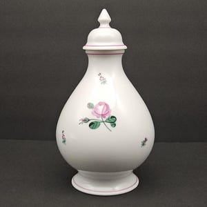 Peut inclure: Un vase en céramique blanc avec un couvercle, orné d'une rose rose et de feuilles vertes. Le vase a un corps bulbeux, une base évasée et un fleuron pointu sur le couvercle. Des bordures roses ornent le bord et la base.