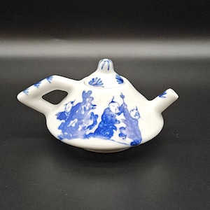 May include: A small white teapot with a blue and white design. The teapot has a lid and a spout. The design features a group of people in traditional clothing.