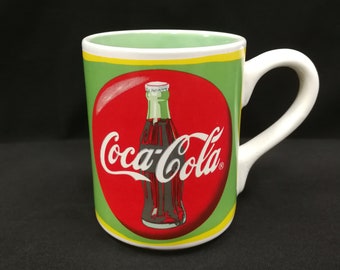 May include: A white ceramic mug with a green and yellow border. The mug features a red circle with a Coca-Cola bottle inside. The bottle is brown and has the Coca-Cola logo on it.