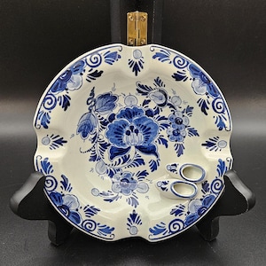 May include: A white ceramic ashtray with a blue and white floral design. The ashtray has a scalloped edge and features two small blue and white shoes.