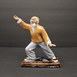 Op de afbeelding: Een keramieken beeldje van een oudere man in een vechtkunstpose. De figuur draagt een mosterdkleurig jasje met witte bies, lichtgrijze broek en zwarte schoenen. Het beeldje is gemonteerd op een rechthoekige basis met bruine en witte tinten.