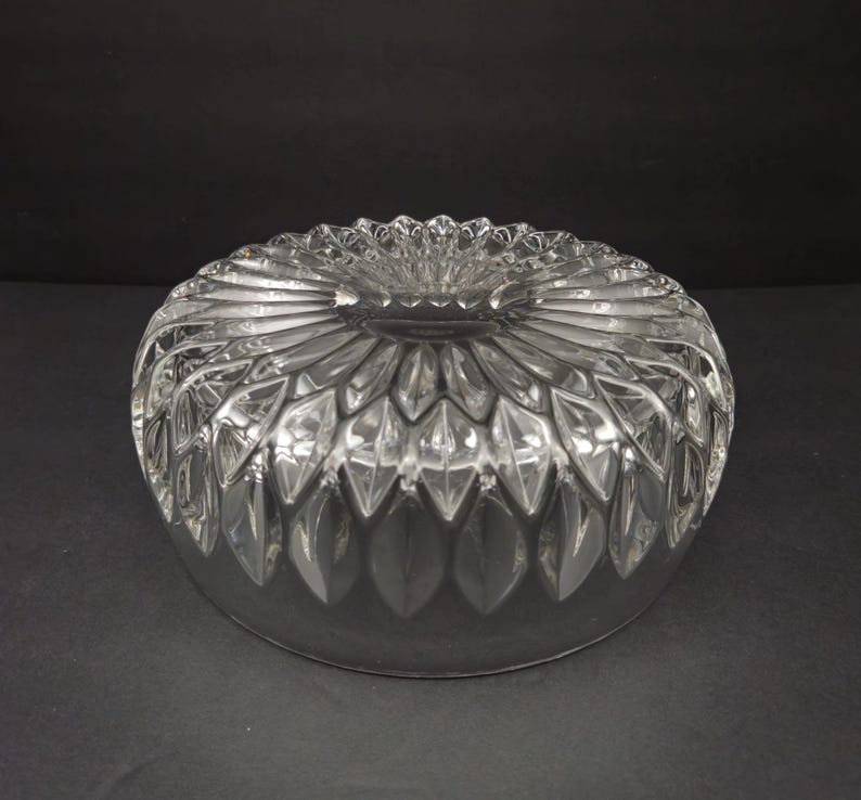 May include: A clear, round glass bowl with a detailed, cut-glass pattern. The bowl features a central, flower-like design surrounded by radiating, leaf-shaped cuts. The rim has a scalloped edge, and the bowl sits on a dark surface.