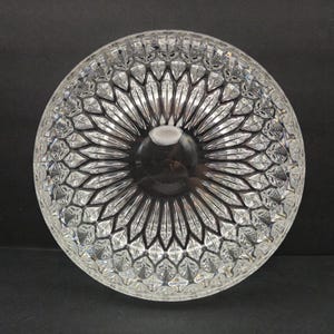 May include: A clear, circular glass bowl with an intricate geometric design. The bowl has a central indentation and radiating lines, forming a starburst pattern. This decorative bowl is likely for serving or display.