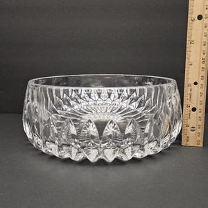 May include: A clear, cut-glass bowl with a decorative pattern. The bowl is approximately 7.6 cm in height and 15.2 cm in diameter. The bowl has a thick base and a detailed, textured design. A wooden ruler is next to the bowl.