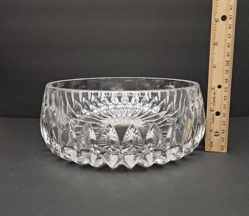 May include: A clear, cut-glass bowl with a decorative pattern. The bowl is approximately 7.6 cm in height and 15.2 cm in diameter. The bowl has a thick base and a detailed, textured design. A wooden ruler is next to the bowl.
