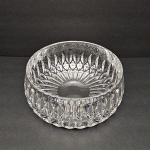 May include: A clear, round crystal bowl with a detailed, cut-glass pattern. The bowl has a wide, shallow design with a textured exterior and interior. The intricate cuts create a sparkling effect, reflecting light. Suitable for serving or display.