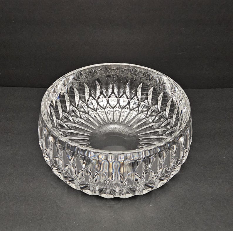 May include: A clear, round crystal bowl with a detailed, cut-glass pattern. The bowl has a wide, shallow design with a textured exterior and interior. The intricate cuts create a sparkling effect, reflecting light. Suitable for serving or display.