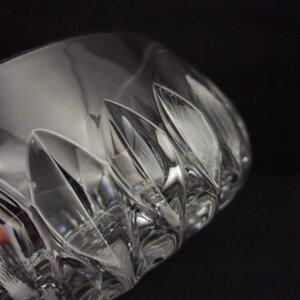 May include: Close-up of a clear, cut-glass drinking glass. The glass has a series of vertical, pointed cuts around the rim, creating a textured surface. The glass is partially visible against a dark background, highlighting its clarity.
