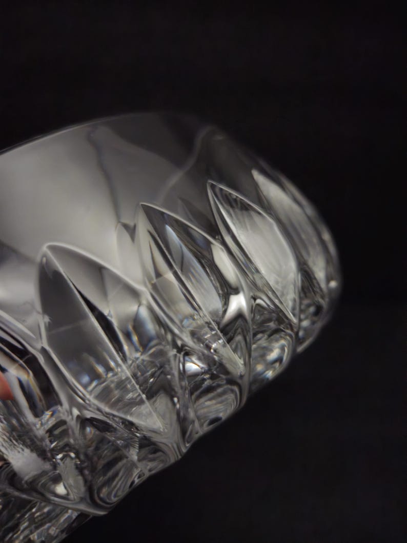 May include: Close-up of a clear, cut-glass drinking glass. The glass has a series of vertical, pointed cuts around the rim, creating a textured surface. The glass is partially visible against a dark background, highlighting its clarity.