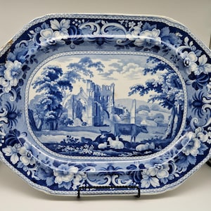 Puede incluir: Una fuente ovalada de cerámica azul y blanca con una escena de paisaje de una ruina de edificio, vacas y ovejas. El borde presenta diseños florales y de volutas. La fuente está sostenida por un soporte de metal negro.