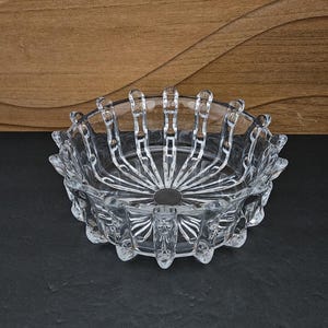 Op de afbeelding: Een heldere, decoratieve glazen kom met een stervormig ontwerp in het midden. De kom heeft een geschulpte rand met verticale, puntige verlengingen. De kom staat op een donkere ondergrond, met een houten achtergrond.