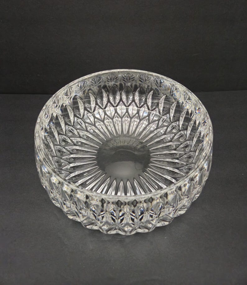 May include: A clear crystal bowl with detailed cut patterns. The bowl has a circular base and radiating lines, creating a starburst effect. The rim features a repeating diamond design. The bowl is approximately 20 cm in diameter.