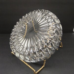 May include: A clear glass bowl with a textured, starburst design, supported by a gold-coloured metal stand. The bowl's pattern consists of repeating, pointed shapes radiating outwards. The stand is made of twisted metal.