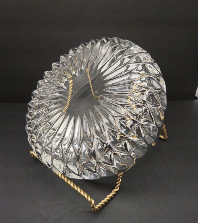 May include: A clear glass bowl with a textured, starburst design, supported by a gold-coloured metal stand. The bowl's pattern consists of repeating, pointed shapes radiating outwards. The stand is made of twisted metal.
